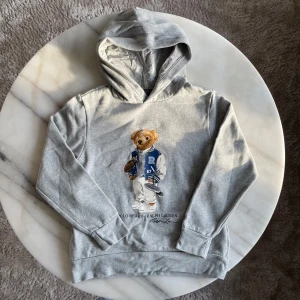 Ralph lauren polo bear - Säljer en grå hoodie från Ralph Lauren med ett coolt björnmotiv på framsidan. Hoodien har långa ärmar och en klassisk huva. Perfekt för en avslappnad stil. 🐻