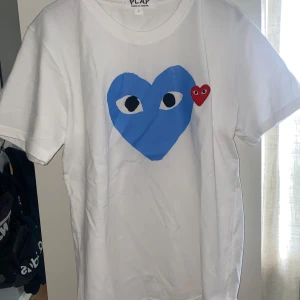 Vit t-shirt från Comme des Garçons - Säljer en vit t-shirt från Comme des Garçons med ett stort blått hjärta med ögon och ett litet rött hjärta på bröstet. T-shirten är kortärmad och har en klassisk rund halsringning. Perfekt för en stilren och lekfull look.