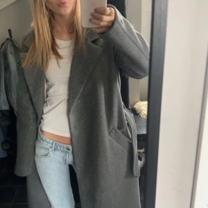 Lång grå kappa - Säljer denna stilrena grå kappa med bälte. Kappan har en klassisk design med långa ärmar och djupa fickor. Relativt ny, köpte/använd i ca 2 veckor, nytt skick, köpte på vero Moda för 650kr, hittar den inte på hemsidan, storlek M men passar bra på S också. Jag är 164❤️