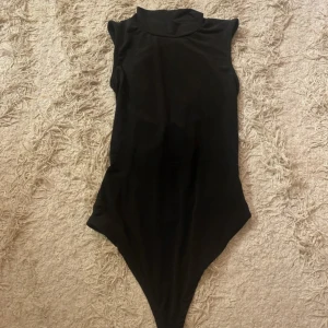 Svart ärmlös bodysuit - Snygg svart ärmlös bodysuit med hög hals och knytning i ryggen. Perfekt för en stilren look.
