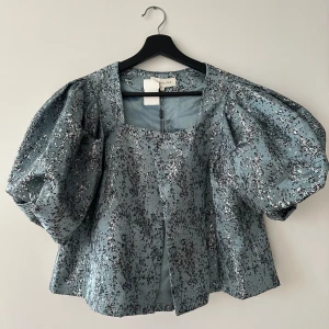 Blå glittrig blus från By Malina - Säljer en elegant blå blus från By Malina med glittrigt mönster. Blusen har puffiga ärmar och en fyrkantig halsringning. Perfekt för att lysa upp vilken outfit som helst!