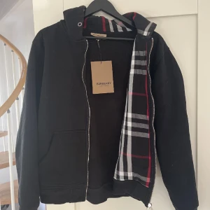 Burberry hoodie  - Säljer en svart hoodie från Burberry med klassiskt rutigt mönster i fodret. Den har en dragkedja framtill och en broderad detalj på bröstet. Perfekt för en stilren look. Finns inget snöre 