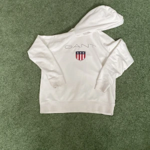Vit hoodie från GANT(storlek s/160) - Säljer en stilren vit hoodie från GANT med ett tryck av deras logotyp och ett emblem på bröstet. Hoodien har långa ärmar och en klassisk huva. Perfekt för en avslappnad stil.