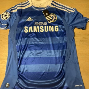 Blå fotbollströja från Adidas - Säljer en blå fotbollströja från Adidas med vit text och detaljer. Tröjan har korta ärmar och ett randigt mönster. Den har tryck med 'Drogba' och nummer 11 på baksidan samt Champions League-märke på ärmen. Perfekt för fotbollsfans!