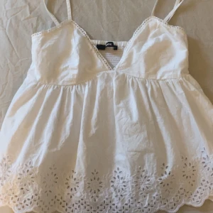 Vit ärmlös topp från BikBok - Säljer en söt vit ärmlös topp från BikBok med spetsdetaljer och broderad nederkant. Toppen har smala axelband och en v-ringad design som ger en romantisk touch. Perfekt för varma dagar!