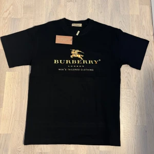 Svart Burberry t-shirt - Skriv dm för bild med tröjan på. Snygg svart t-shirt från Burberry med deras ikoniska logga i guld på bröstet. Tillverkad i mjuk bomull för en bekväm passform. Perfekt för dig som vill ha en stilren look med en touch av lyx. Storlek ”S” (men lite stor i storlek)