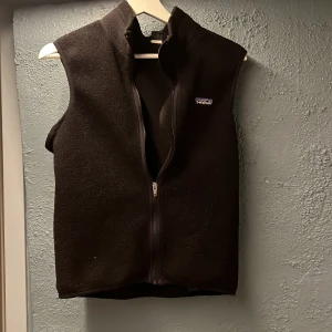 Svart fleeceväst från Patagonia - Säljer säljer en svin fett retro  svart fleeceväst från Patagonia som är perfekt nu till våren. Västen har en enkel och stilren design, perfekt för lager-på-lager under kyligare dagar. Den har en liten logga på bröstet och en praktisk ficka på baksidan.