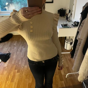 Beige ribbad tröja med knappar - En beige ribbad tröja med dekorativa knappar framtill och på ärmarna. Tröjan har långa ärmar och en jätte najs passform som framhäver kroppen bra. Säljer då den inte kommer till användning 