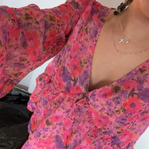 Blommig blus med volangdetaljer - Säljer en färgglad blommig blus med volangdetaljer och v-ringning. Blusen har långa ärmar och en lös passform, perfekt för en somrig look. Den har en mix av rosa, lila och röda nyanser. Passar perfekt till sommaren såsom midsommar🥰