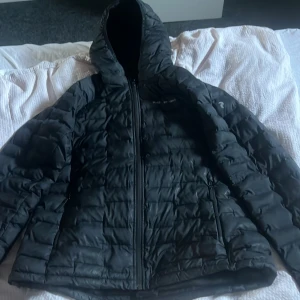 Svart dunjacka från Peak Performance - Säljer en svart dunjacka från Peak Performance. Jackan har en dragkedja framtill och en huva för extra skydd mot kyla. Perfekt för kalla dagar med sin quiltade design som håller värmen inne. En stilren och funktionell jacka för vintern.