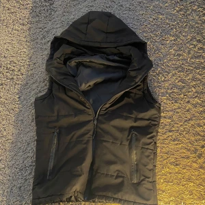 Svart väst - Snygg svart dunväst med dragkedja och praktiska fickor framtill. Västen har en huva som ger extra skydd mot väder och vind. Perfekt för kyligare dagar. Priset kan diskuteras 