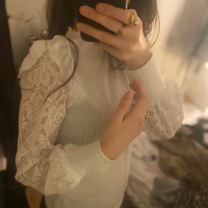 Vit ribbad topp med spetsdetaljer - Säljer en elegant vit ribbad topp med spetsdetaljer på ärmarna. Toppen har långa ärmar och en hög krage som ger en stilren look. Perfekt för att lyfta vilken outfit som helst.