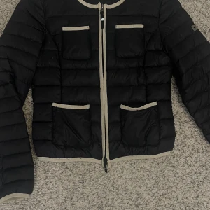 Svart dunjacka från Twin Set - Snygg svart dunjacka från Twin Set med beige detaljer. Jackan har en dragkedja framtill och fyra fickor med beige kant. Perfekt för kyligare dagar.