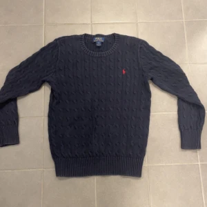 Mörkblå stickad tröja från Ralph Lauren - Säljer en snygg mörkblå stickad tröja från Ralph Lauren med kabelstickat mönster. Tröjan har en rund halsringning och det klassiska röda logotypbroderiet på bröstet. Perfekt för kyligare dagar!