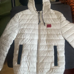 Vit quiltad jacka från Hugo - Säljer en  vit quiltad jacka från Hugo med svart foder och dragkedja. Jackan har en huva med snörning och praktiska fickor. Perfekt för kyligare dagar.