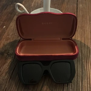 Svarta solglasögon från Gucci - Snygga svarta solglasögon från Gucci med en fyrkantig form. De har en elegant design med guldiga GG-logotyper på sidorna. Perfekta för att ge en lyxig touch till din stil.