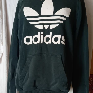 Grön hoodie från Adidas - Säljer en grön hoodie från Adidas med det klassiska trefoil-logotypen i vitt på framsidan. Tröjan har en stor ficka framtill och långa ärmar. Perfekt för en avslappnad stil. Hör av dig vid intresse!