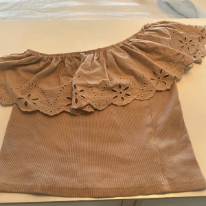 Beige offshoulder topp från Zara - Säljer en söt beige offshoulder topp från Zara med vackra spetsdetaljer. Toppen har en ribbad struktur och en volangkant som ger en romantisk touch. Perfekt för varma sommardagar!