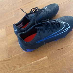 Svarta fotbollsskor från Nike Phantom GT - Säljer ett par svarta Nike Phantom GT fotbollsskor med blå detaljer och snörning. Skorna har en modern design med en tydlig logga och är perfekta för fotbollsplanen. Innersulan är röd och ger en snygg kontrast.