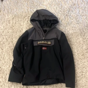 Svart och grå jacka från Napapijri - Säljer en stilren svart och grå jacka från Napapijri med huva och en stor ficka framtill. Jackan har en broderad logotyp och norska flaggan på bröstet. 