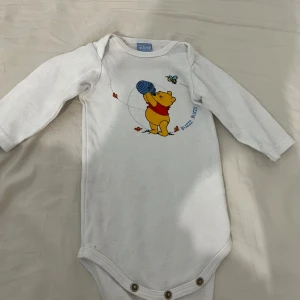 Vit långärmad body med Nalle Puh-motiv - Söt vit långärmad body från Disney med ett färgglatt Nalle Puh-motiv på framsidan. Perfekt för små äventyrare! Bodyn har praktiska knappar i grenen för enkel på- och avklädning.
