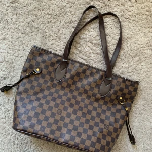 Brun rutig axelväska - Säljer en snygg brun axelväska med klassiskt rutigt mönster. Väskan har långa handtag och en rymlig insida med röd foder. Något sliten vid remmarna som syns på bild men annars inget fel på den! 