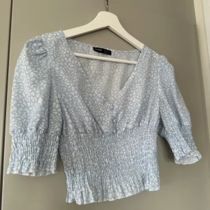 Ljusblå blus - Säljer en söt ljusblå blus med vita leopardmönster. Blusen har en v-ringad design och korta puffärmar med smockdetaljer. Perfekt för en somrig look!