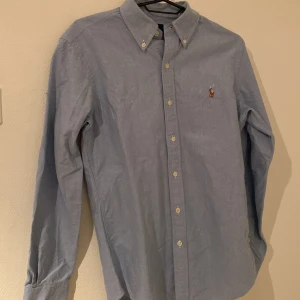Blå skjorta från Ralph Lauren - Säljer en stilren blå skjorta från Ralph Lauren med klassisk krage och knappar framtill. Skjortan har en broderad logotyp på bröstet och långa ärmar. Perfekt för en snygg och avslappnad look. Använd 2 gånger köpt för 1599 kan gå ner till 500 Max vid snabb affär 