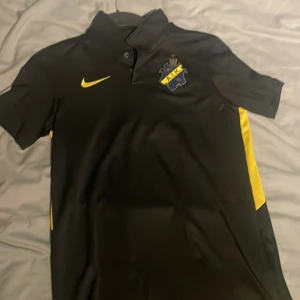 Svart AIK fotbollströja från Nike - Säljer en svart AIK fotbollströja från Nike med gula detaljer och klubbens emblem på bröstet. Tröjan har korta ärmar och en klassisk krage.