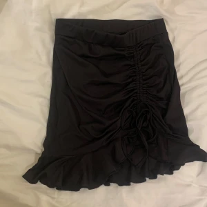 Svart kjol med volang och snörning - Snygg svart kjol med volangdetaljer och snörning framtill. Kjolen har en kort längd, är kortare framtill och är perfekt för sommaren. 