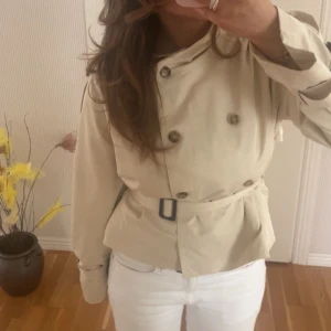 Beige trenchcoat med bälte - Snygg beige trenchcoat med knappar och bälte i midjan. Perfekt för vår och höst. Klassisk design med dubbelknäppning och stilren look.
