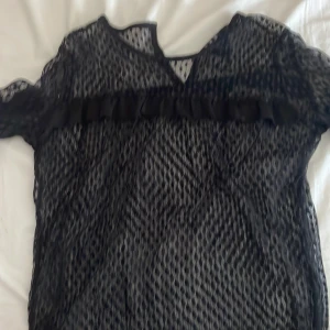 Svart spetsblus från ONLY - Snygg svart spetsblus från ONLY med volangdetaljer. Blusen har korta ärmar och ett transparent mönster som ger en elegant look. Perfekt för att piffa upp vilken outfit som helst!