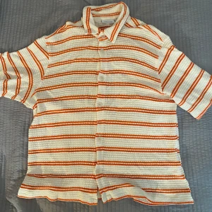 Randig kortärmad linneskjorta från Weekday - Säljer en snygg randig linneskjorta från Weekday i vitt och orange. Skjortan är kortärmad och har knappar framtill. Perfekt för en avslappnad stil med en unik textur.