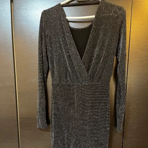 Svart glittrig klänning - Snygg svart klänning med glittrigt material och v-ringad design. Klänningen har långa ärmar och en elegant passform som passar perfekt för festliga tillfällen.