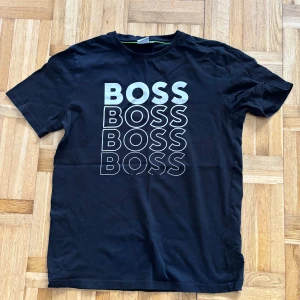 Svart t-shirt från Boss - Säljer en svart t-shirt från Boss med texten 'BOSS' tryckt flera gånger på framsidan. T-shirten har en slim fit passform och är kortärmad. Perfekt för en stilren och avslappnad look.