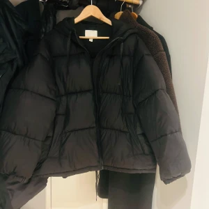 Svart pufferjacka med huva - Säljer en svart pufferjacka med huva. Jackan har en dragkedja framtill och är perfekt för kyligare dagar. Den har en oversized passform som ger en avslappnad look. Bär vanligtvis xs och denna jacka är i storlek M och sitter därför oversize på mig