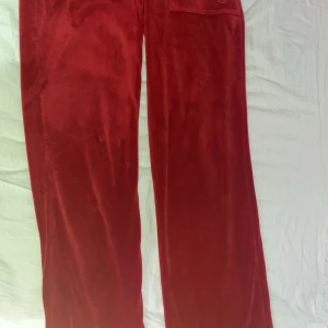 Röda mjukisbyxor från Juicy Couture - Snygga röda mjukisbyxor från Juicy Couture i en mjuk och glansig finish. Byxorna har en bekväm resårmidja med snörning och en ficka bak. Perfekta för en avslappnad stil.