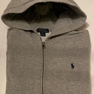 Grå hoodie från Polo Ralph Lauren - Säljer en stilren, ny grå hoodie från Polo Ralph Lauren med dragkedja och broderad logga på bröstet. Perfekt för en avslappnad look. Hoodien har en bekväm passform.