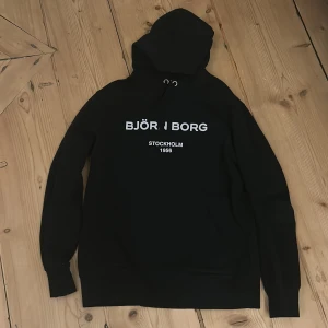 Svart hoodie från Björn Borg - Säljer en svart hoodie från Björn Borg med texten 'Stockholm 1956' tryckt på framsidan. Tröjan har en klassisk design med huva och långa ärmar. Perfekt för en avslappnad stil.