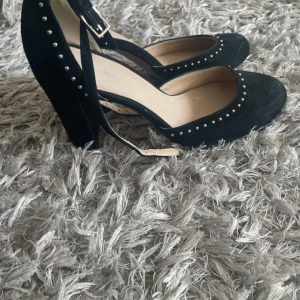 Svarta pumps med nitar fårn pier one - Snygga svarta pumps i mocka med rund tå och klack. Skorna har dekorativa nitar längs kanterna och ett justerbart spänne runt ankeln. Perfekta för att ge en edgy touch till din outfit. Från pier one.