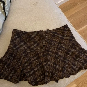 Brun rutig kjol med knappar - Snygg brun rutig kjol med knappar framtill. Kjolen har en volangdetalj vid nederkanten som ger en fin touch. Perfekt för en trendig look! Mycket bra skick