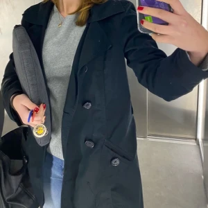 Svart kappa med knappar - Snygg svart kappa med dubbelknäppning och klassisk krage. Perfekt för kyligare dagar. Jackan har en stilren design med fickor framtill.