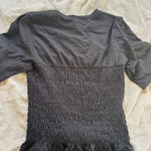 Svart topp från Divided - Säljer en svart topp från Divided med en smockad design på kroppen och trekvartsärmar. Toppen har en rund halsringning och en volangdetalj vid nederkanten. Perfekt för en stilren look!