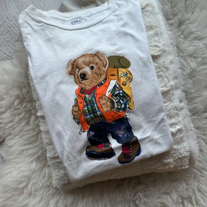 Vit t-shirt med björntryck från Polo Ralph Lauren - Säljer en vit t-shirt från Polo Ralph Lauren med björntrycket. Avslappnad passform, köpt från barnavdelningen (XL), men är en M på vuxen. 🧸