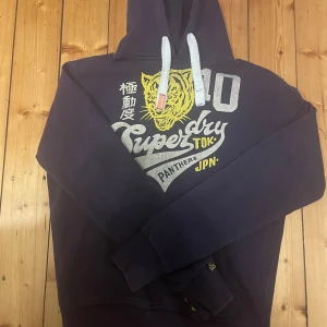 Mörkblå hoodie från Superdry - Säljer en mörkblå hoodie från Superdry med en stor gul och vit tigerdesign och text på framsidan. Hoodien har en justerbar huva med vita snören och ribbade muddar vid ärmslut och nederkant. Perfekt för en avslappnad stil!
