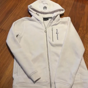Vit hoodie från Sail Racing storlek 150, 200kr - Säljer en vit hoodie från Sail Racing med dragkedja och huva. Den har en logotyp på bröstet och en liten ficka med dragkedja. Perfekt för en avslappnad stil.