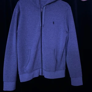 Mörkgrå Ralph Lauren zip - Snygg grå hoodie från Ralph Lauren med dragkedja och klassisk logga på bröstet. Perfekt för en avslappnad stil. Den har långa ärmar och en bekväm passform.
