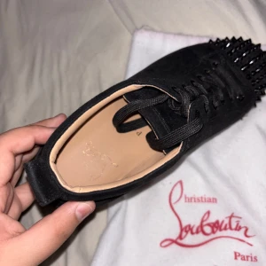 Svarta sneakers med nitar från Louboutin - Snygga svarta sneakers från Christian Louboutin med nitar på tån. Skorna har en klassisk design med snörning och en röd sula som ger en ikonisk touch. Perfekta för att ge din outfit en edgy stil.