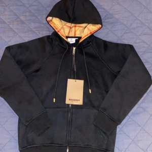 Svart hoodie från Burberry - Säljer en stilren svart hoodie från Burberry med dragkedja och justerbar huva. Insidan av huvan har det klassiska Burberry-mönstret i beige och rött. Perfekt för en avslappnad och trendig look. Står S men sitter mer som barn storlek L(A-cop)