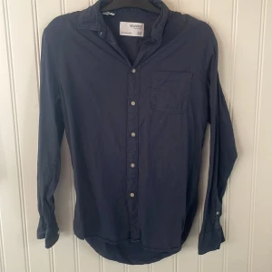 Mörkblå skjorta från Selected Homme - Snygg mörkblå skjorta från Selected Homme med klassisk krage och vita knappar framtill. Skjortan har en bröstficka och långa ärmar med knappar vid ärmsluten. Perfekt nu till sommaren.